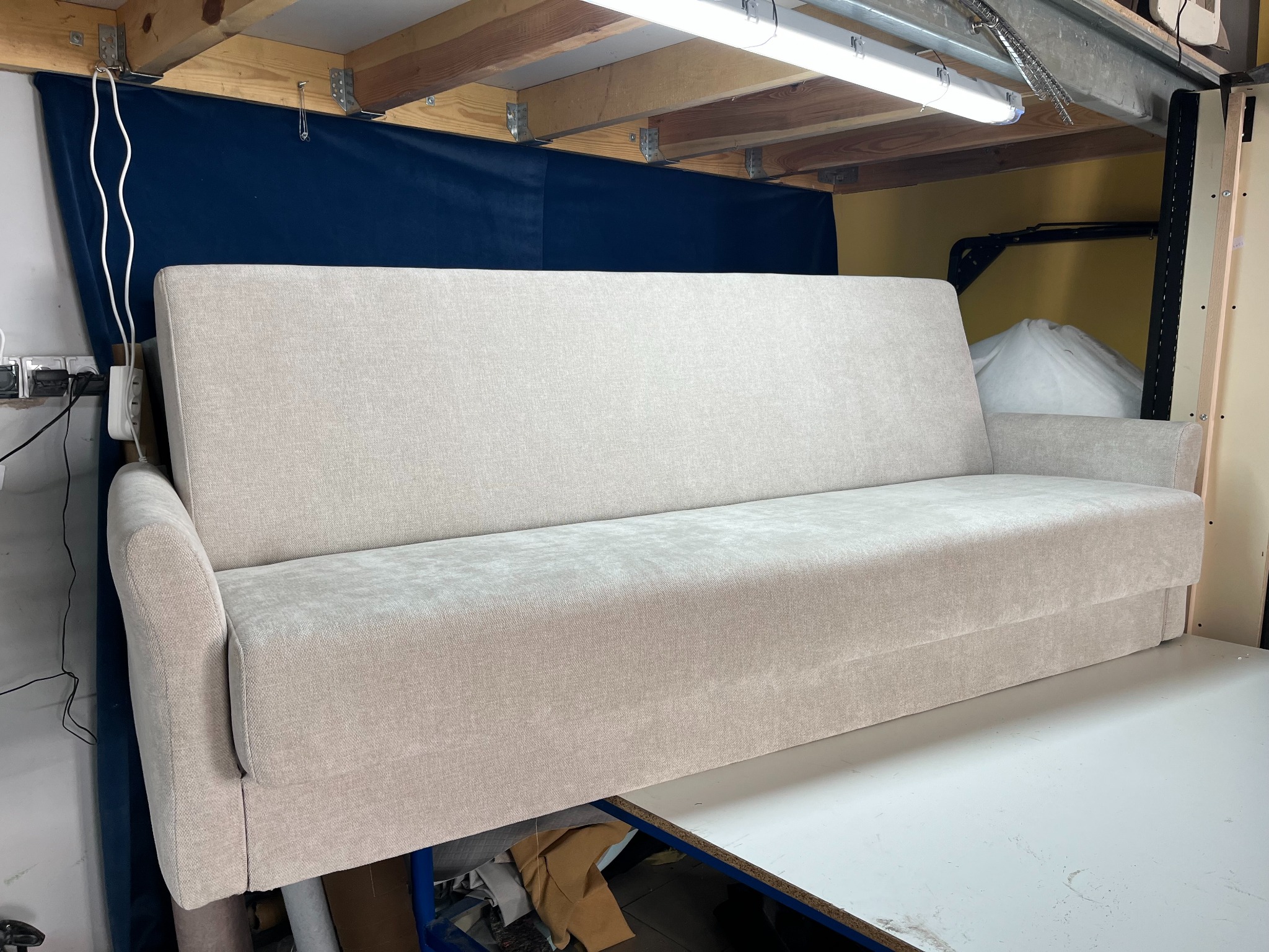 Jasna, tapicerowana sofa rozkładana na stole w warsztacie, z widoczną drewnianą konstrukcją sufitu i fragmentem niebieskiego materiału w tle.