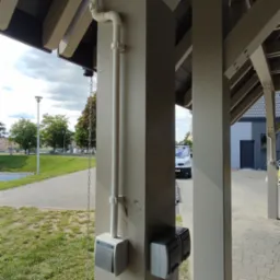 Zewnętrzna instalacja elektryczna w peszlu, zamontowana na drewnianej konstrukcji z szarymi gniazdkami. Widok na zewnątrz budynku, chodnik i zieleń.