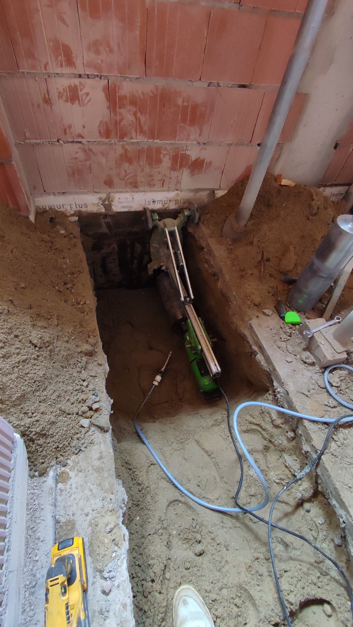 Wykop z zieloną wiertnicą rdzeniową, przewody, ceglany strop i fragment białego buta. Praca w ziemi z użyciem specjalistycznego sprzętu wewnątrz budynku.