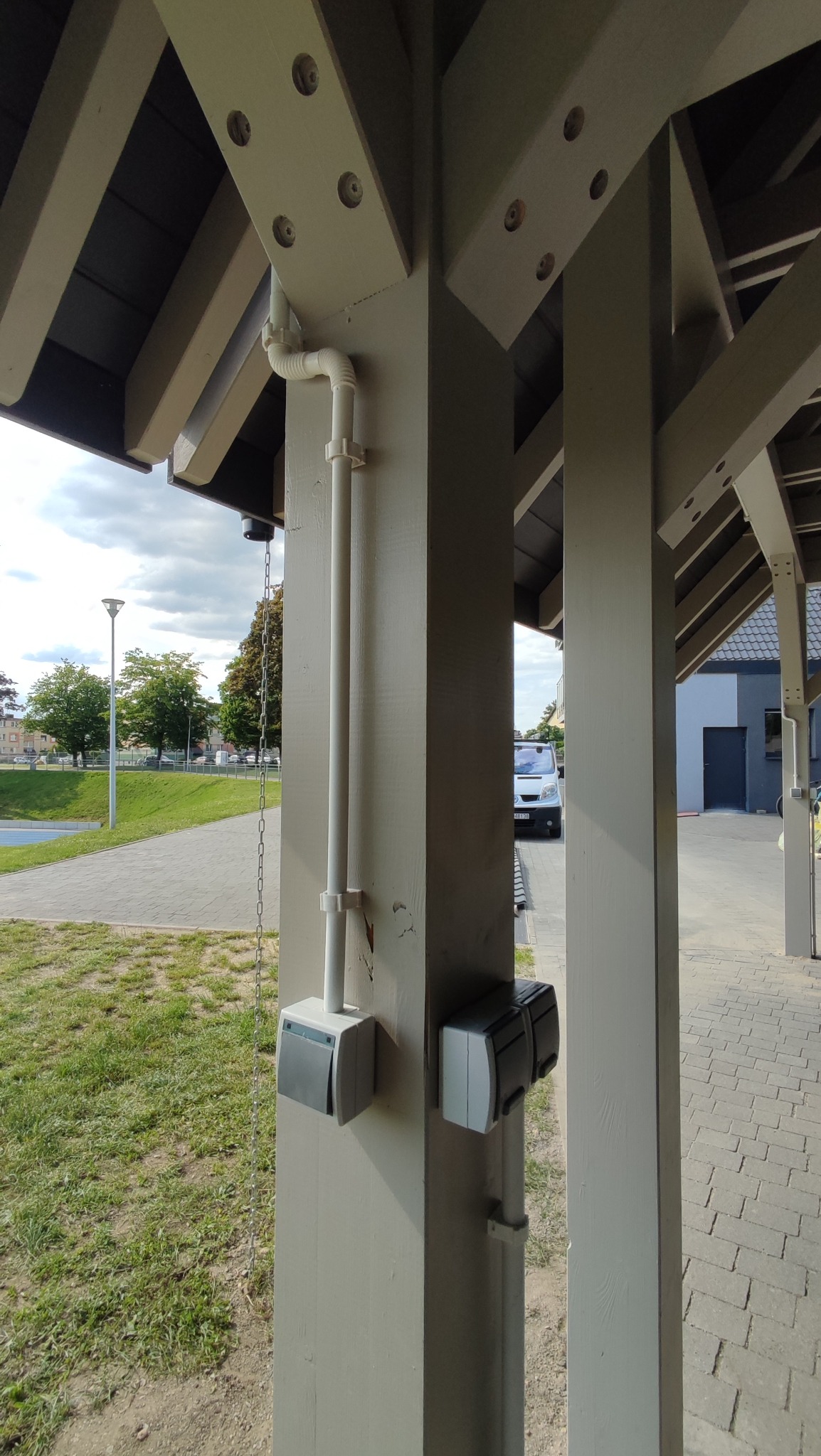 Zewnętrzna instalacja elektryczna w peszlu, zamontowana na drewnianej konstrukcji z szarymi gniazdkami. Widok na zewnątrz budynku, chodnik i zieleń.