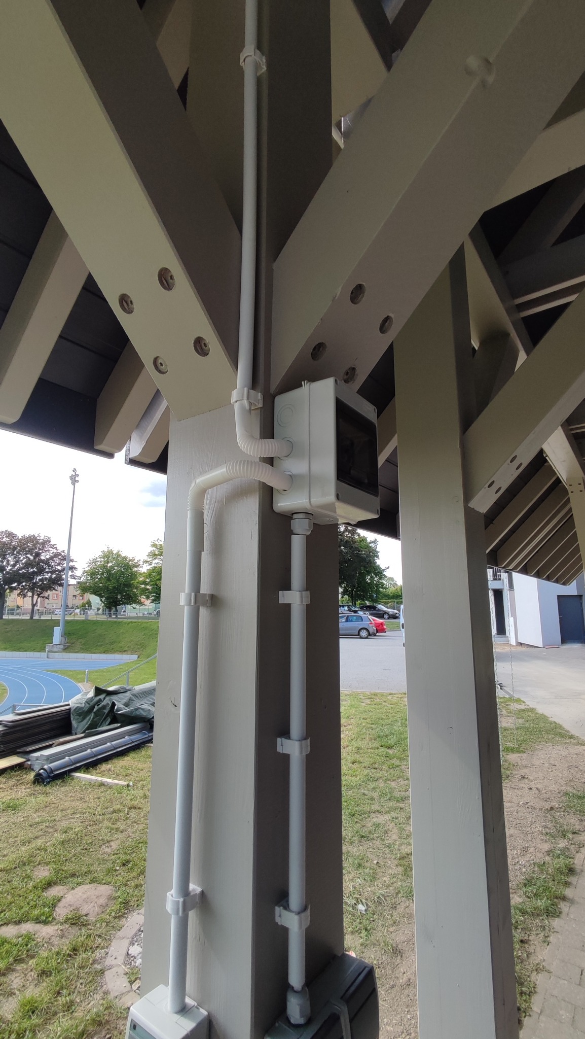 Biała skrzynka elektryczna z rurami PCV, zamontowana na drewnianej konstrukcji w zadaszonym miejscu. Widoczne elementy mocujące rury i fragment otoczenia.