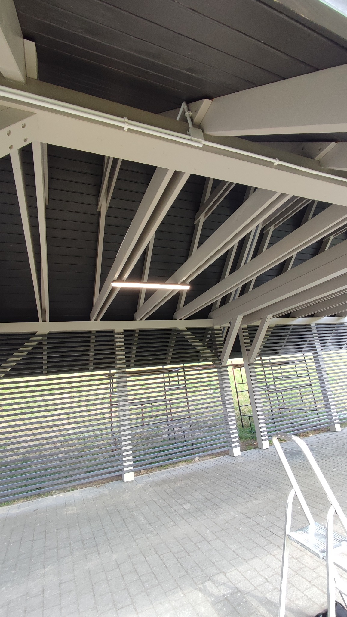 Oświetlenie LED pod stropem zadaszonej konstrukcji z drewnianymi belkami i ażurowymi ścianami. Widoczne białe rury instalacji elektrycznej.