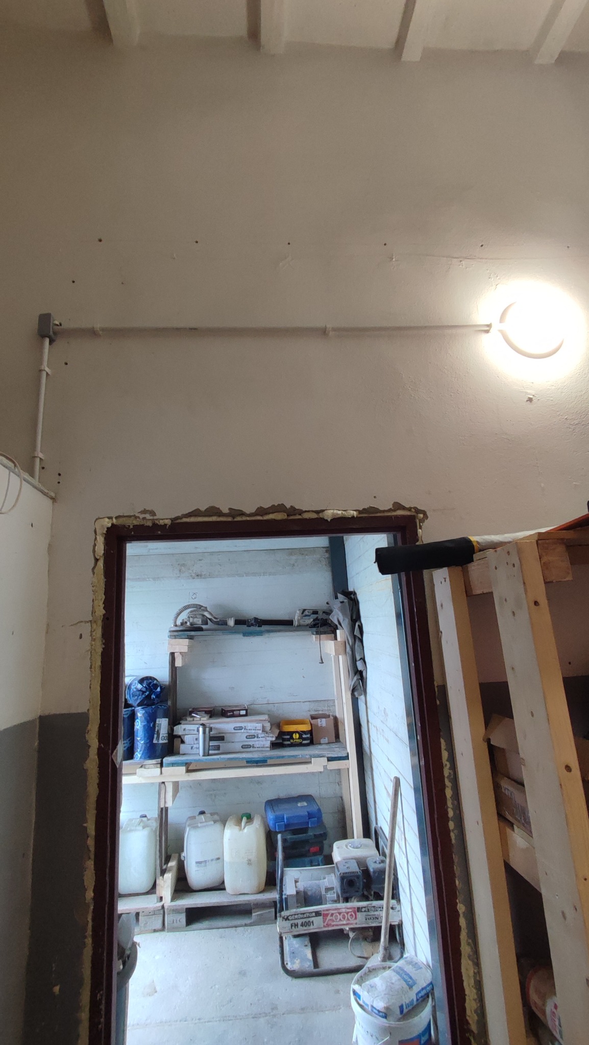 Wnętrze z instalacją elektryczną: przewód w peszlu pod sufitem i lampa. W tle regał z narzędziami i materiałami budowlanymi, widoczny przez otwór w ścianie.
