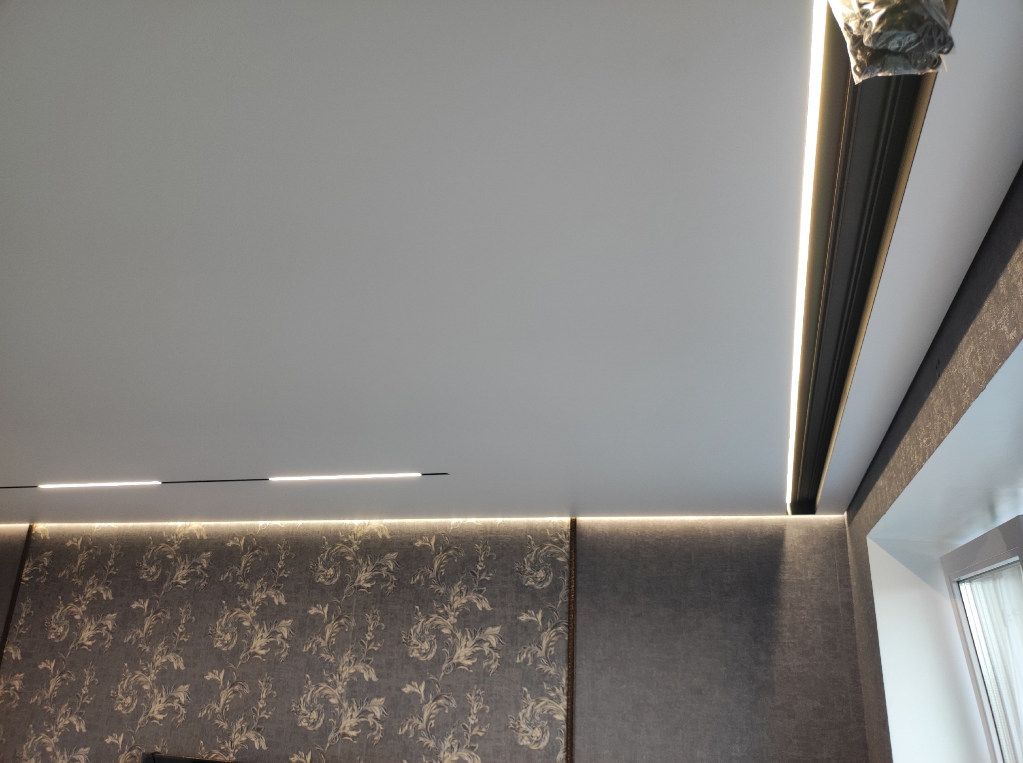 Biały sufit z oświetleniem LED, dekoracyjna ściana z wzorzystą tapetą i szarą lamperią, oraz fragment okna w rogu pokoju. Nowoczesne wykończenie wnętrza.