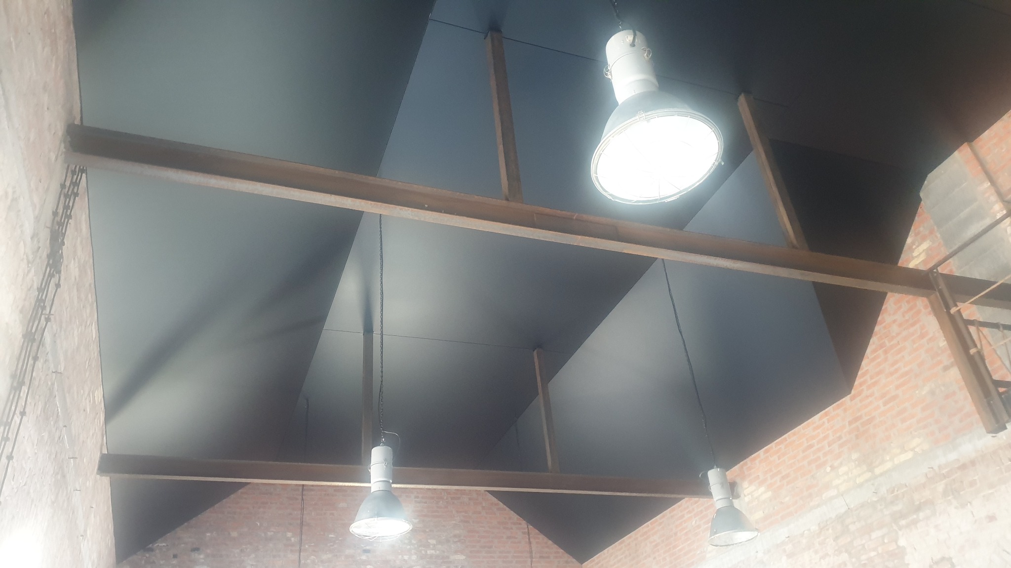 Czarny, geometryczny sufit podwieszany z widocznymi belkami stalowymi i lampami przemysłowymi. Ceglana ściana w tle dodaje surowego charakteru.