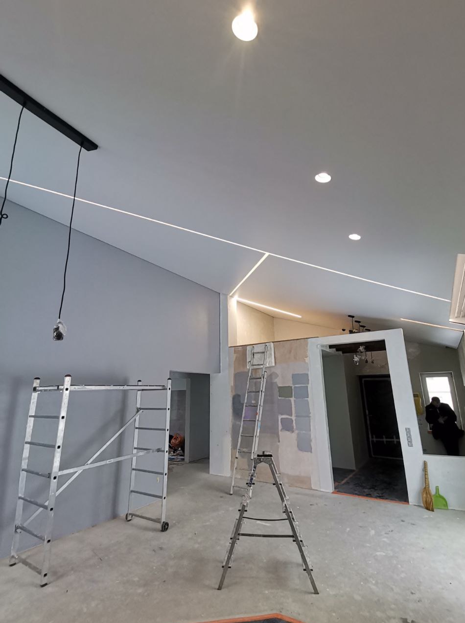 Montaż oświetlenia liniowego w nowym, jasnym wnętrzu ze skośnym sufitem. Widoczne drabiny i osoba siedząca w oknie. Etap wykończeniowy.
