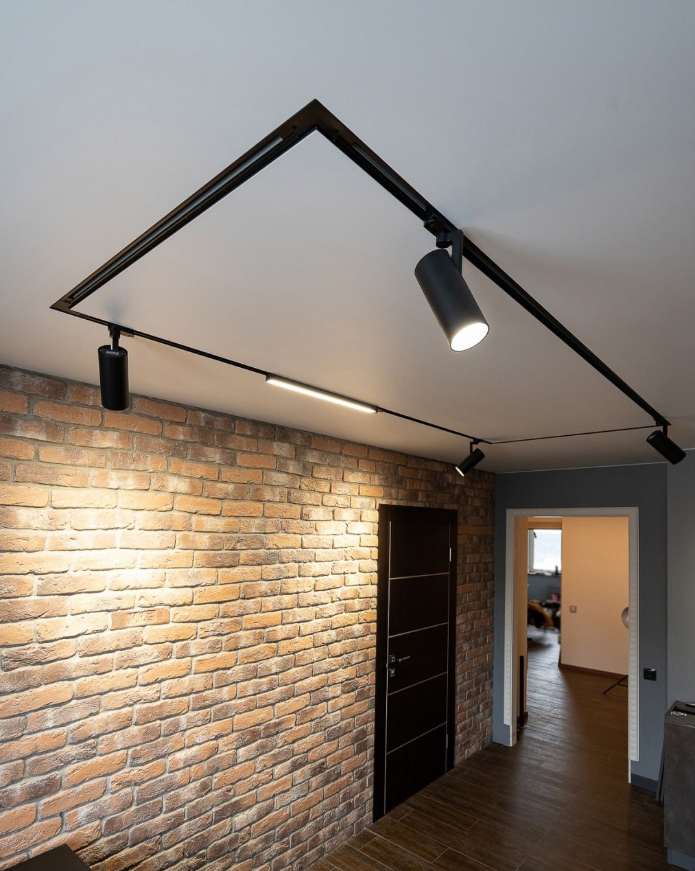 Nowoczesny system oświetlenia szynowego z reflektorami, zamontowany na białym suficie w korytarzu z ceglaną ścianą i czarnymi drzwiami.