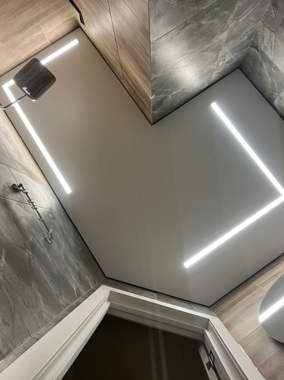 Nowoczesna łazienka z białym, podwieszanym sufitem i liniowym oświetleniem LED, marmurowe płytki ścienne i kabina prysznicowa z deszczownicą.