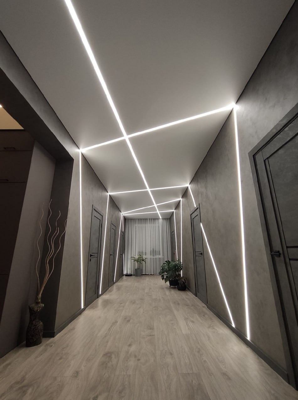 Korytarz z szarymi ścianami i podłogą, oświetlony napinanym sufitem z geometrycznym wzorem z linii świetlnych. Rośliny doniczkowe w tle, dekoracyjny wazon po lewej.