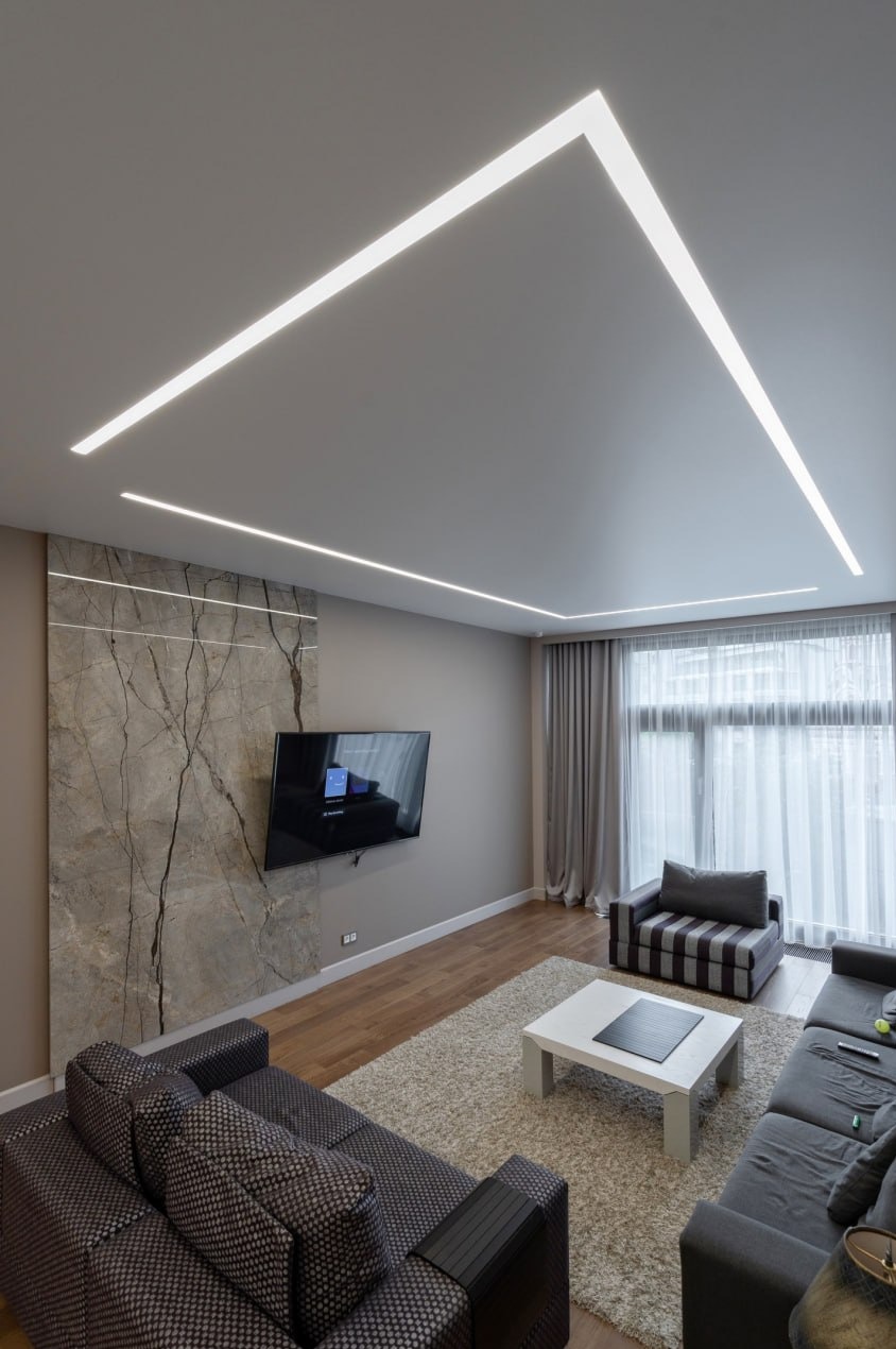 Nowoczesny salon z podświetlanym sufitem: liniowe lampy LED tworzą geometryczny wzór. Ściana z imitacją marmuru, telewizor, szara sofa i dywan.