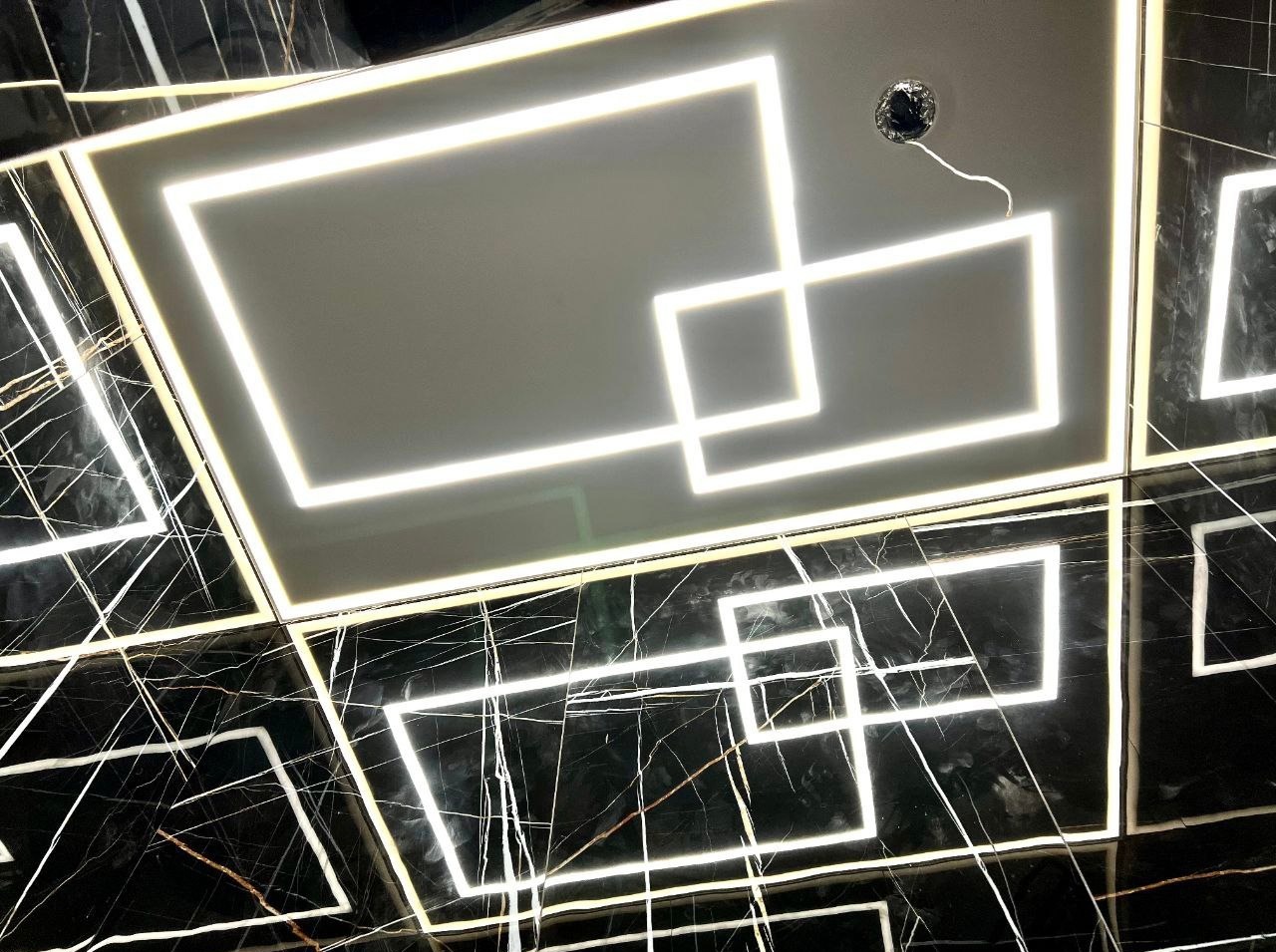 Nowoczesny sufit z podświetleniem LED w kształcie kwadratów, na tle czarnej marmurowej podłogi. Oświetlenie tworzy geometryczny wzór.