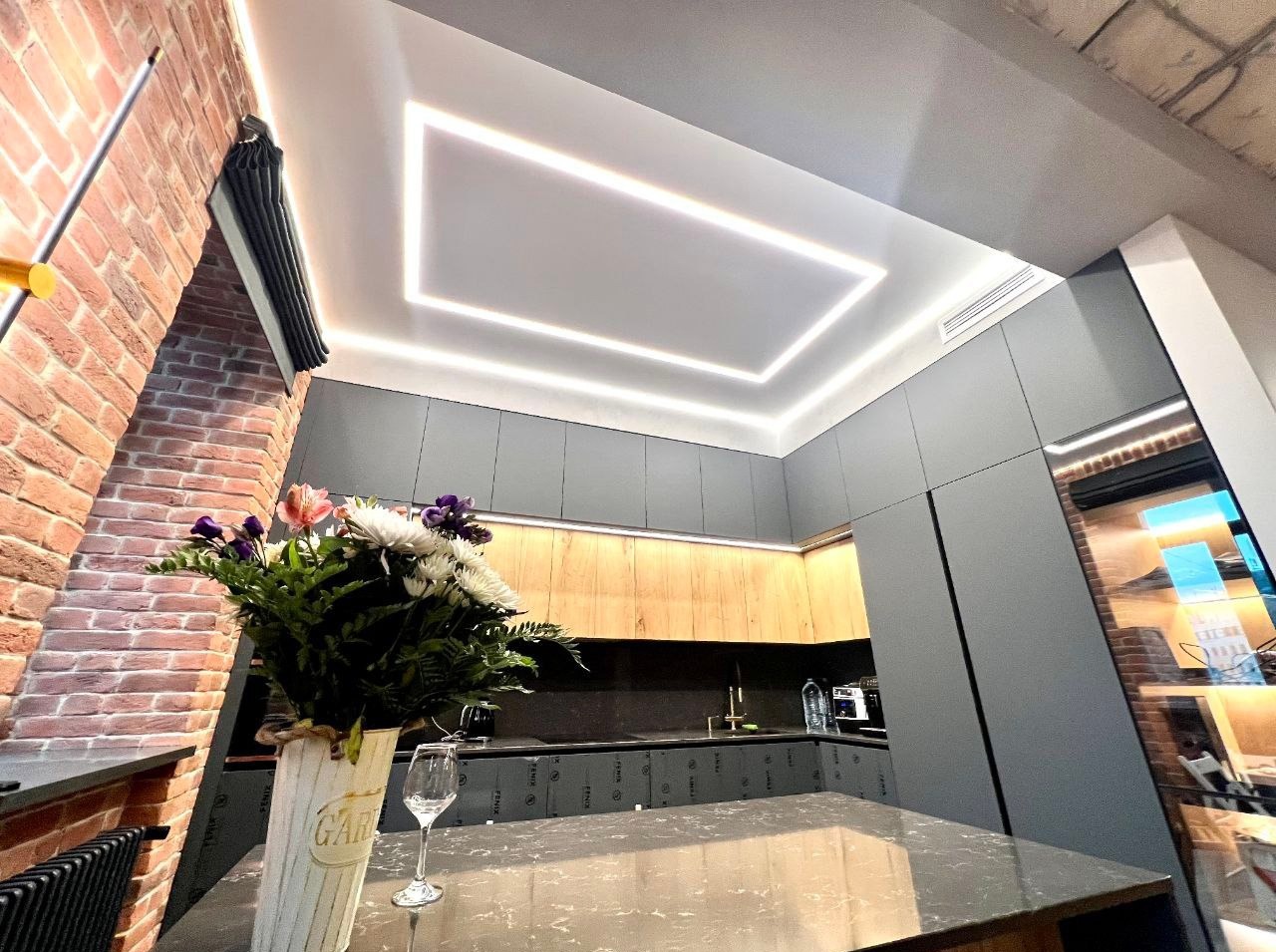 Nowoczesna kuchnia z oświetleniem LED w suficie napinanym tworzącym ramę. Ceglana ściana z dekoracją i bukiet kwiatów na blacie. Szare szafki.