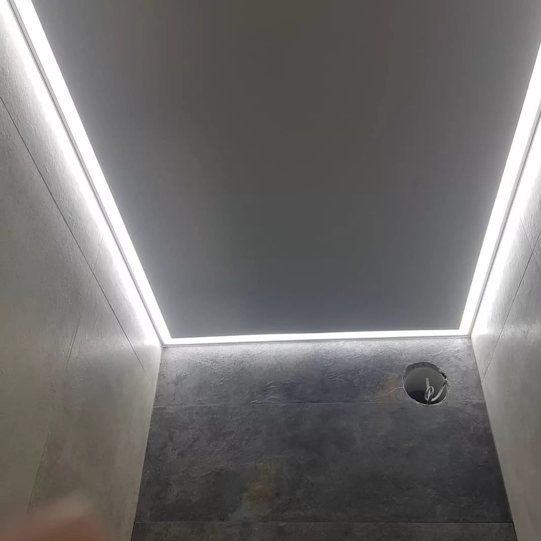 Szary sufit napinany z oświetleniem LED wzdłuż krawędzi, widok z dołu. Na ścianie widoczny otwór instalacyjny z wystającymi przewodami.