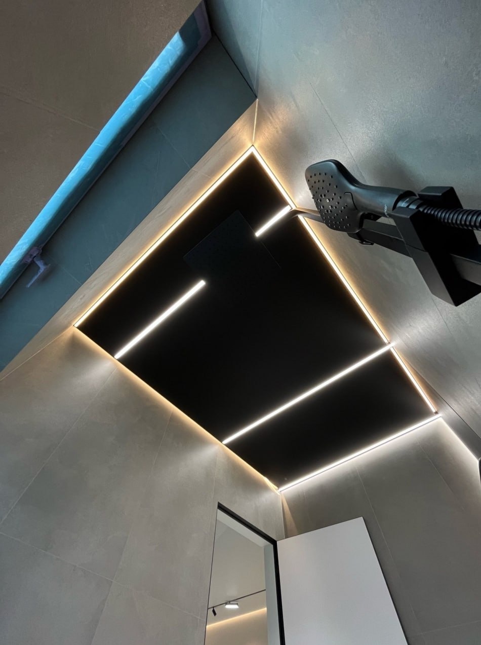 Nowoczesna łazienka z czarnym sufitem i oświetleniem LED wzdłuż krawędzi. Widoczna kabina prysznicowa i szara ściana z płytek. Minimalistyczny design.