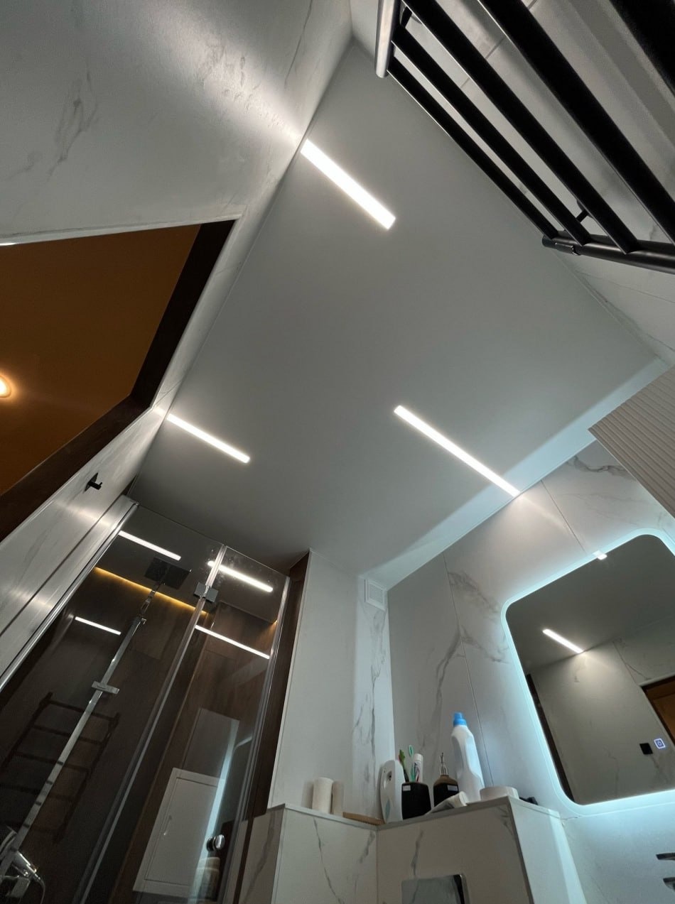 Nowoczesna łazienka z białym sufitem i liniowym oświetleniem LED. Widoczna kabina prysznicowa i podświetlane lustro. Ujęcie z dołu podkreśla geometrię wnętrza.