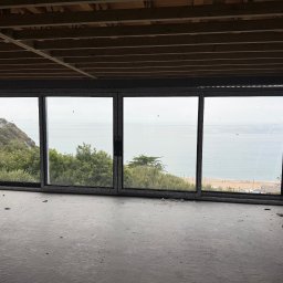 JR MONTER - Czteroskrzydłowe okno przesuwne z widokiem na morze i plażę w surowym wnętrzu. Widoczna drewniana konstrukcja stropu i fragmenty ścian w stanie deweloperskim.