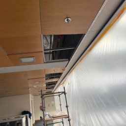 JR MONTER - Biuro w trakcie remontu: widok na sufit z drewnianymi panelami, częściowo odkrytą instalacją i zabezpieczoną folią ścianą. Widoczne rusztowanie.