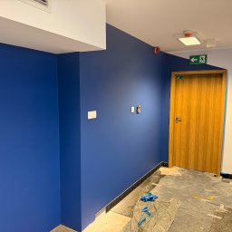 JR MONTER - Niebieska ściana w biurze po malowaniu, widoczne zabezpieczone podłoże, drewniane drzwi z zielonym znakiem ewakuacyjnym i fragment schodów.