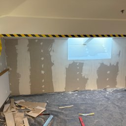 JR MONTER - Remontowane wnętrze z zabezpieczoną podłogą, częściowo wyszpachlowaną ścianą z oknem i taśmą ostrzegawczą. Widoczne narzędzia i płyty gipsowe.