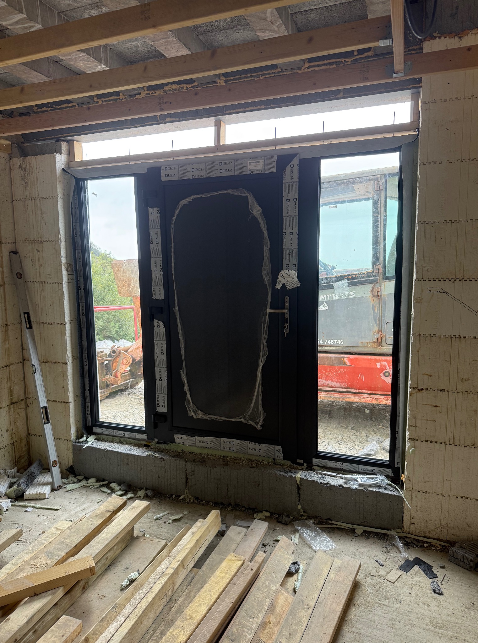 Ciemne drzwi wejściowe z szybą, częściowo zabezpieczone folią, zamontowane w surowym otworze budynku z betonu komórkowego. Na zewnątrz widoczny sprzęt budowlany.