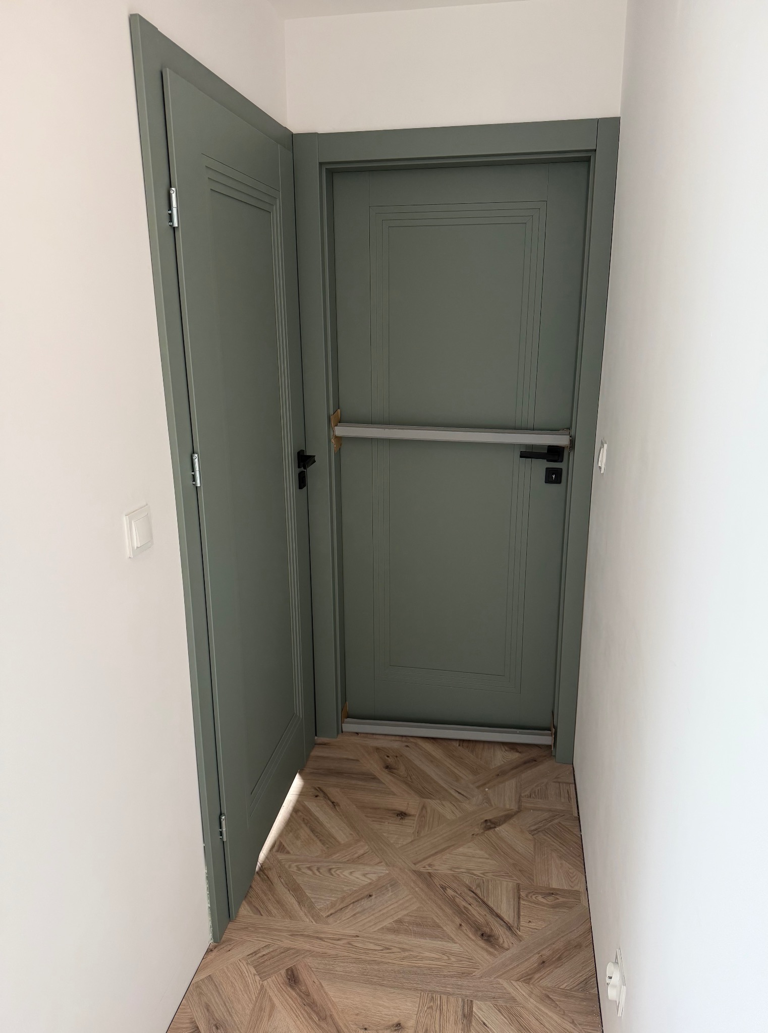 Szara, dwuskrzydłowa brama przeciwpożarowa z poziomą listwą zabezpieczającą, zamontowana w odnowionym wnętrzu z parkietem w jodełkę.