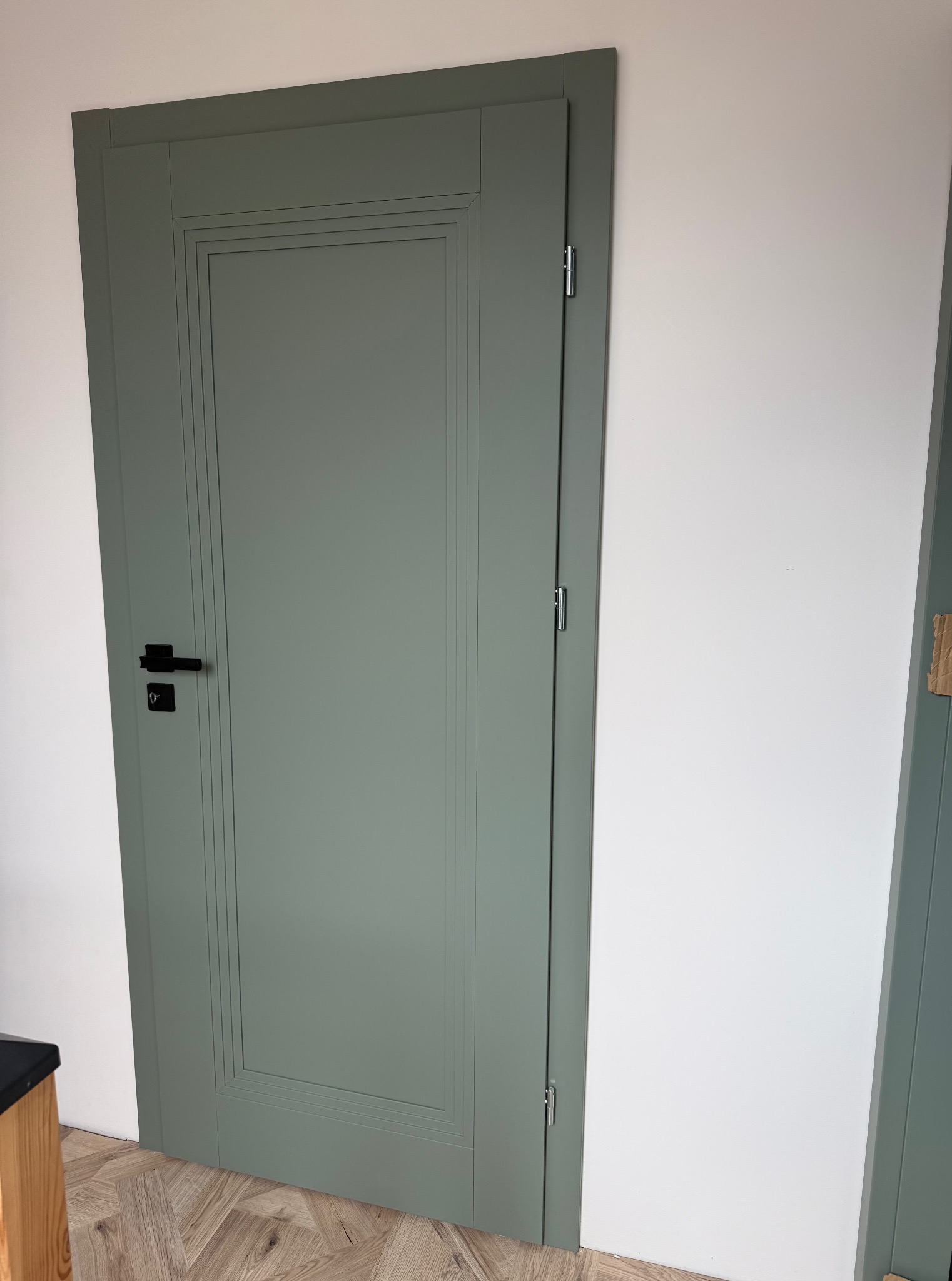 Szare, panelowe drzwi wewnętrzne z czarną klamką, zamontowane w białej ścianie. Podłoga z parkietu w jodełkę. Widoczne zawiasy i fragment drugiej ościeżnicy.