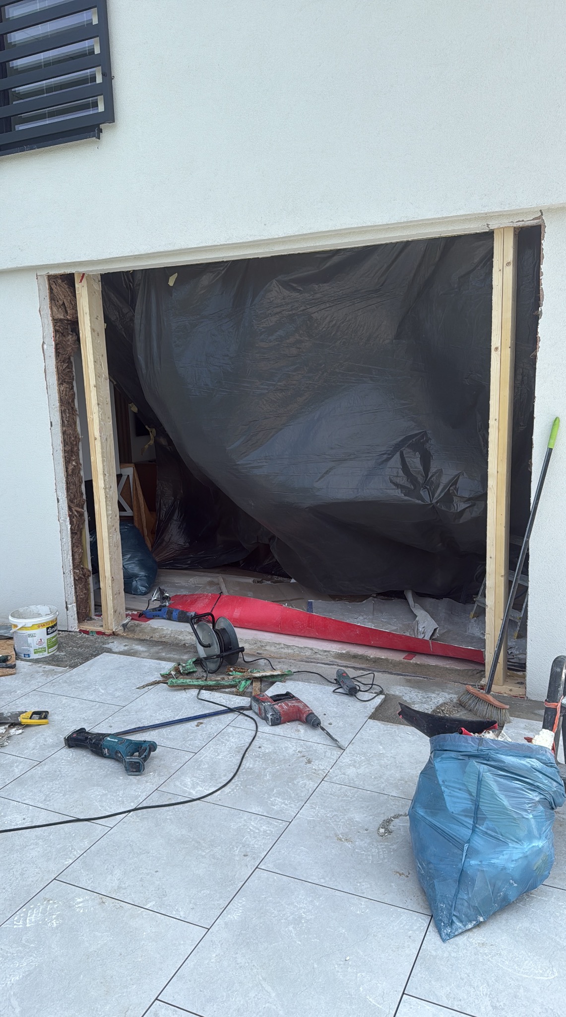 Przygotowanie do montażu drzwi tarasowych: wycięty otwór w ścianie, zabezpieczony folią, narzędzia na płytkach. Widoczne drewniane ramy i ślady prac remontowych.