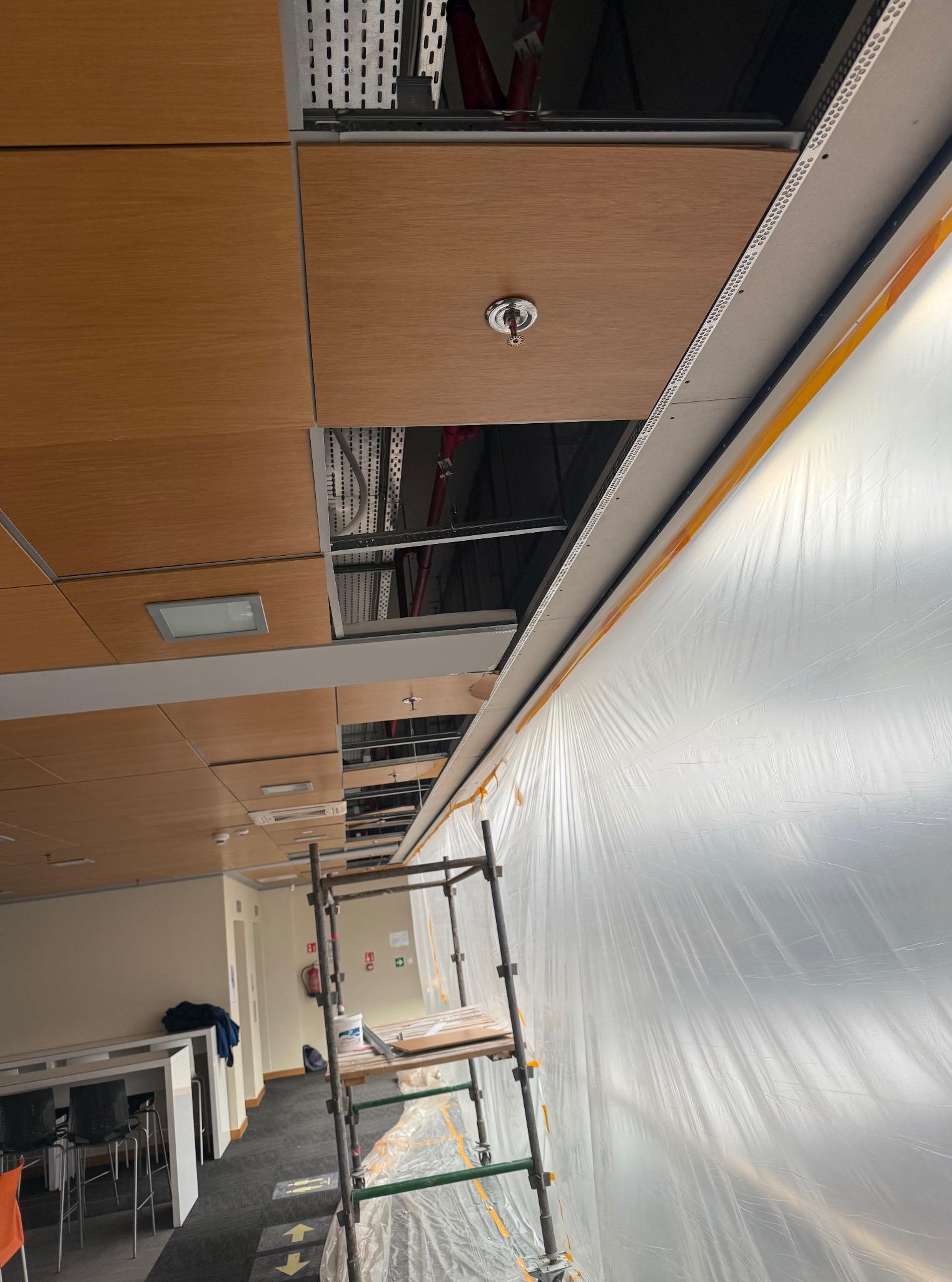 Biuro w trakcie remontu: widok na sufit z drewnianymi panelami, częściowo odkrytą instalacją i zabezpieczoną folią ścianą. Widoczne rusztowanie.