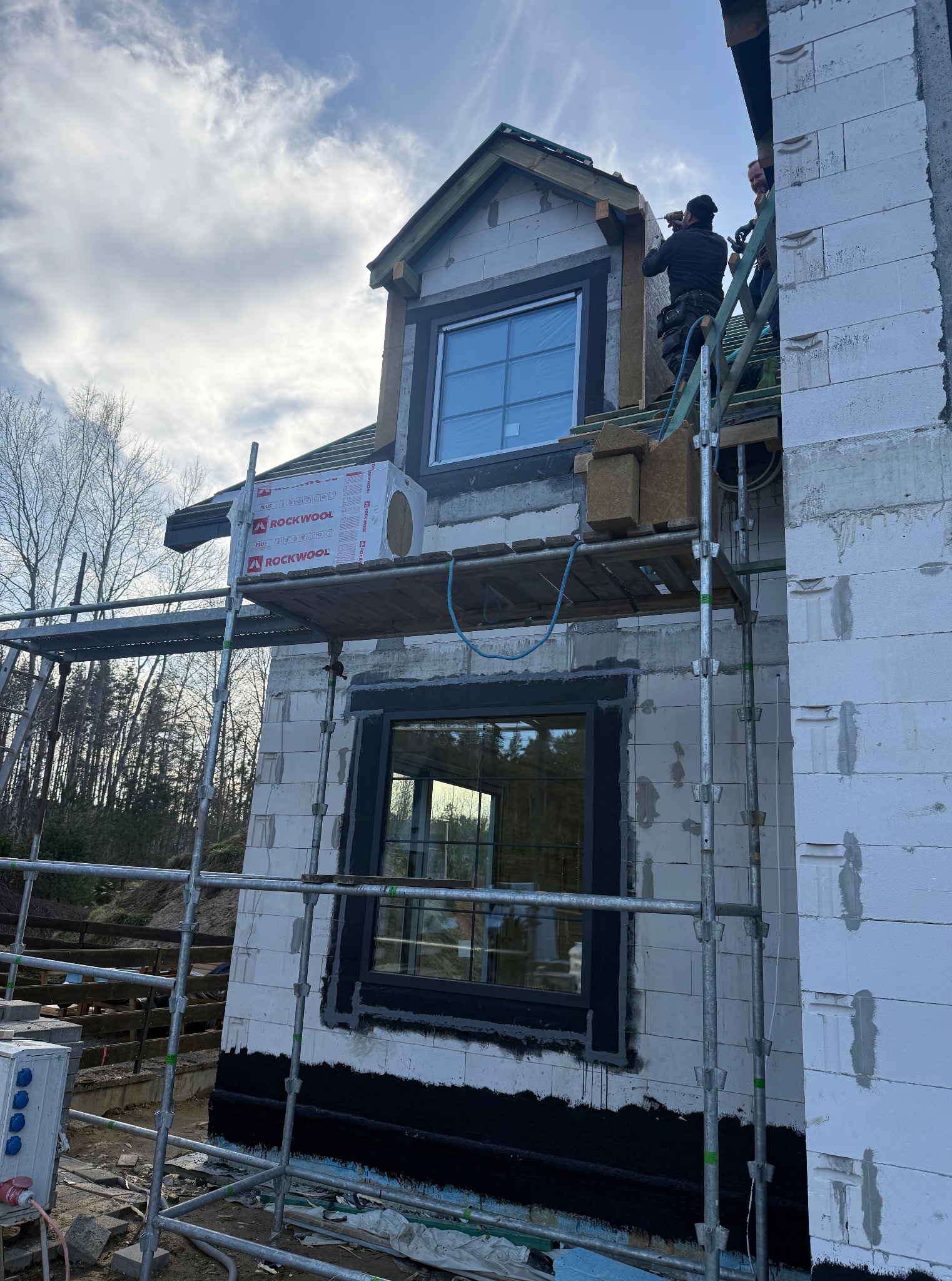 Budowa domu z białych bloczków: pracownicy na rusztowaniu montują okna i ocieplenie Rockwool. Widok z dołu na surowy stan budynku.