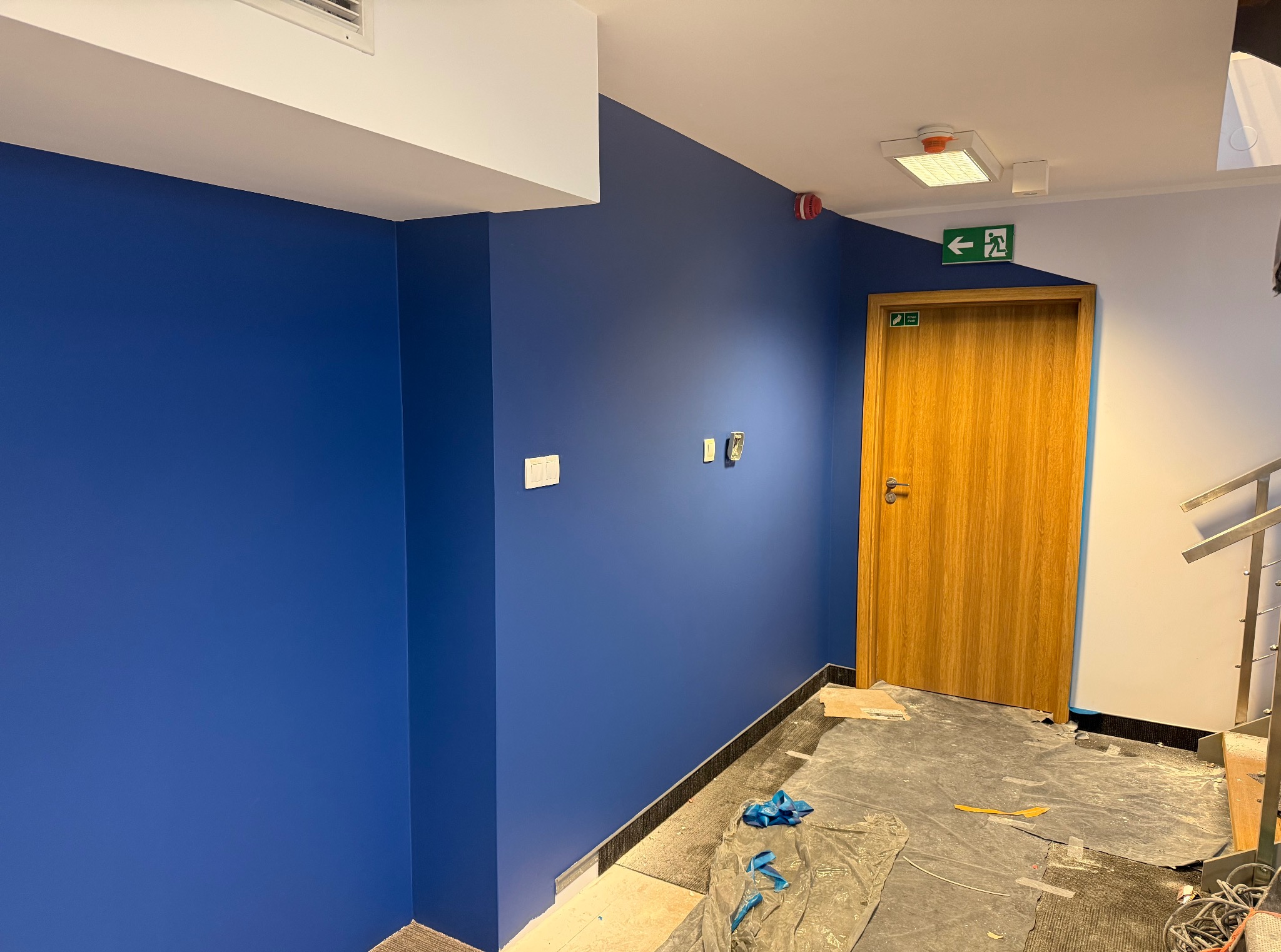 Niebieska ściana w biurze po malowaniu, widoczne zabezpieczone podłoże, drewniane drzwi z zielonym znakiem ewakuacyjnym i fragment schodów.