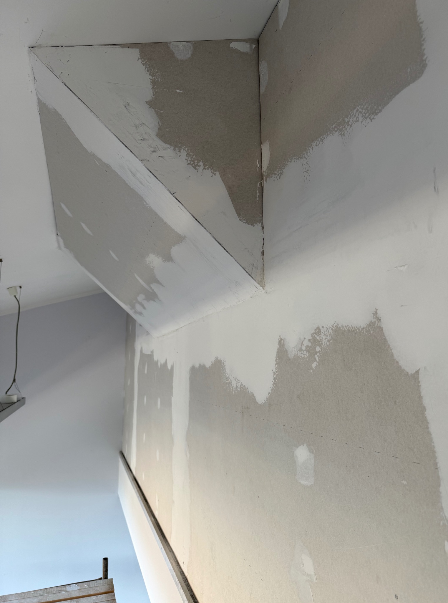 Narożnik ściany i sufitu po szpachlowaniu, widoczne ślady po łatach gipsowych. Na ścianie widoczne pionowe linie pomocnicze.