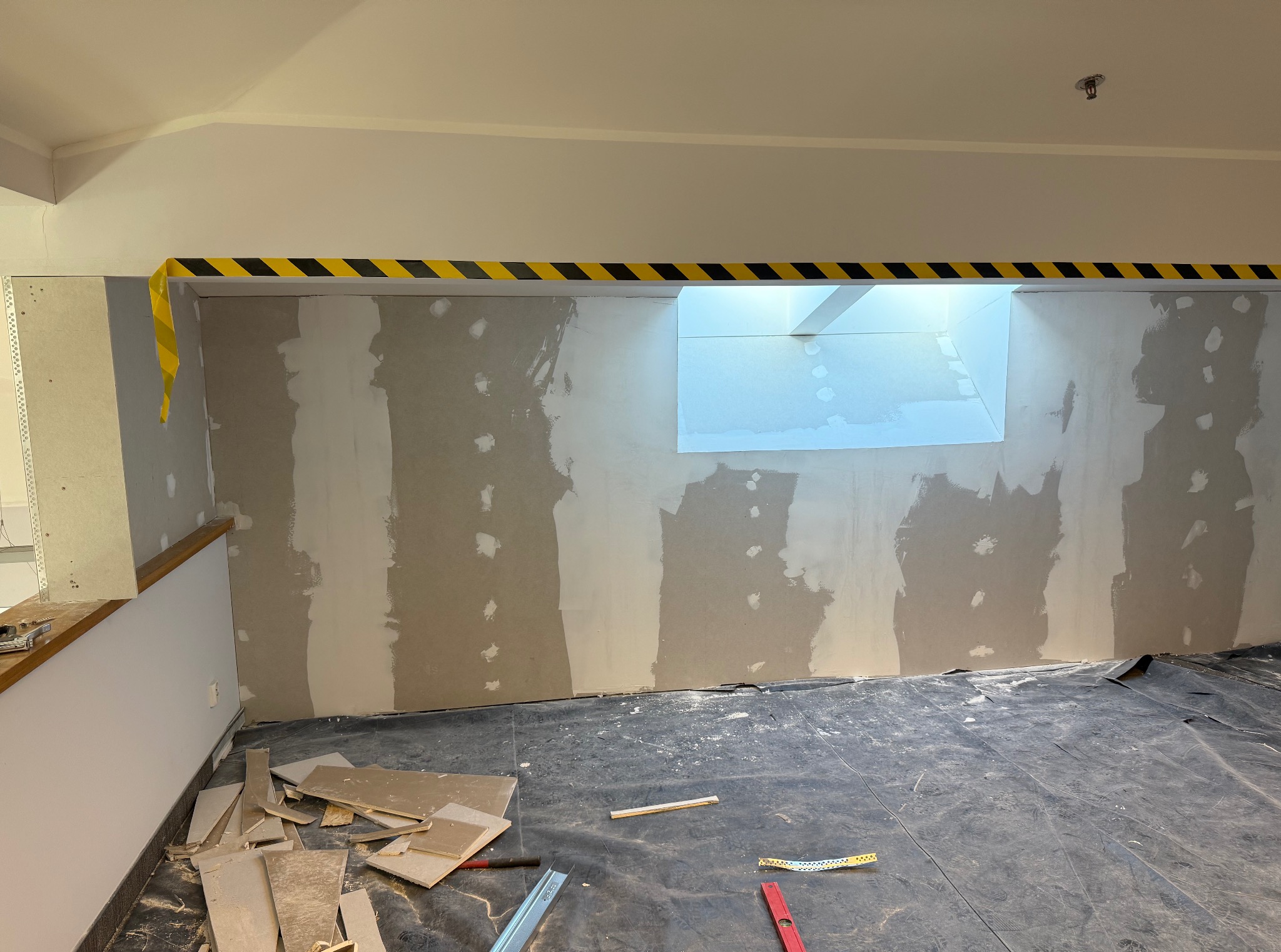 Remontowane wnętrze z zabezpieczoną podłogą, częściowo wyszpachlowaną ścianą z oknem i taśmą ostrzegawczą. Widoczne narzędzia i płyty gipsowe.