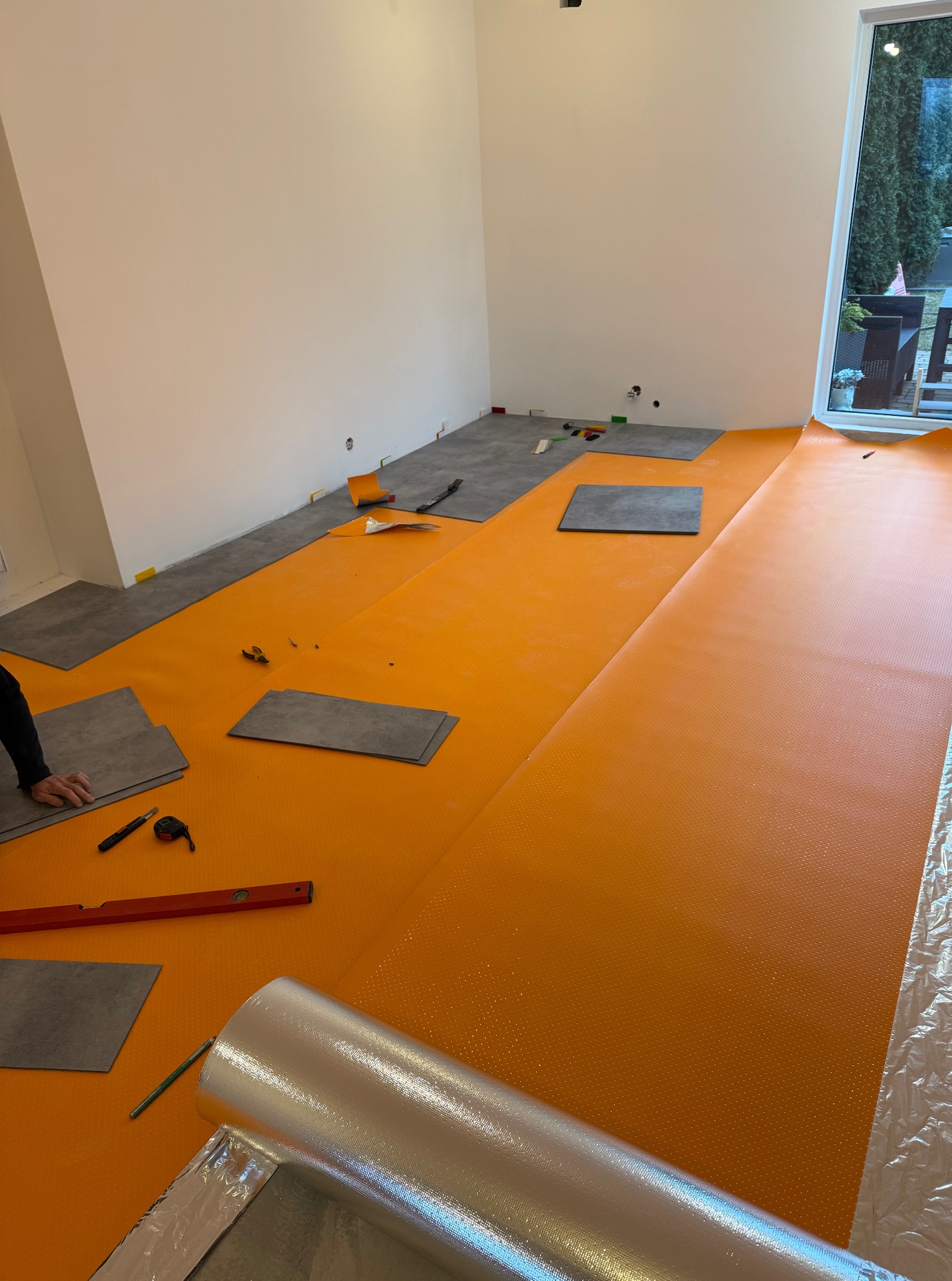 Wnętrze z pomarańczową folią izolacyjną i częściowo ułożonymi panelami podłogowymi. Widoczne narzędzia: poziomica, nóż, miarka. Praca w toku, przygotowanie do finalizacji.