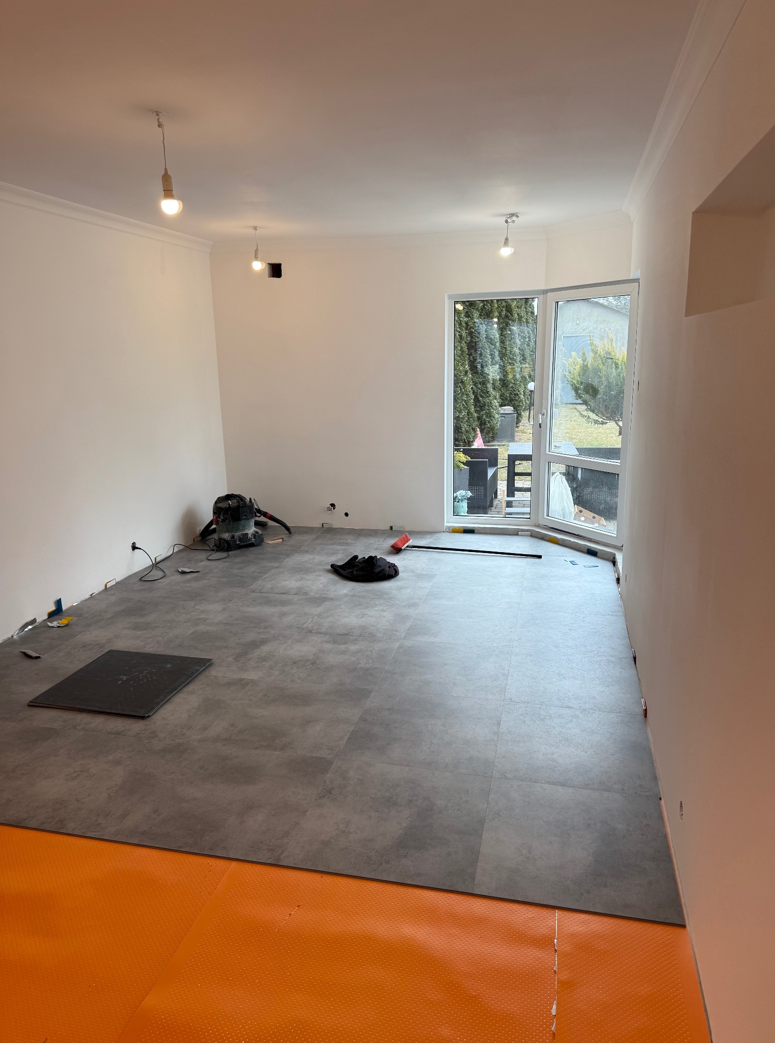 Układanie szarych paneli na pomarańczowej macie w jasnym pokoju z oknem na ogród. Widoczne narzędzia i odkurzacz. Częściowo ułożona podłoga.