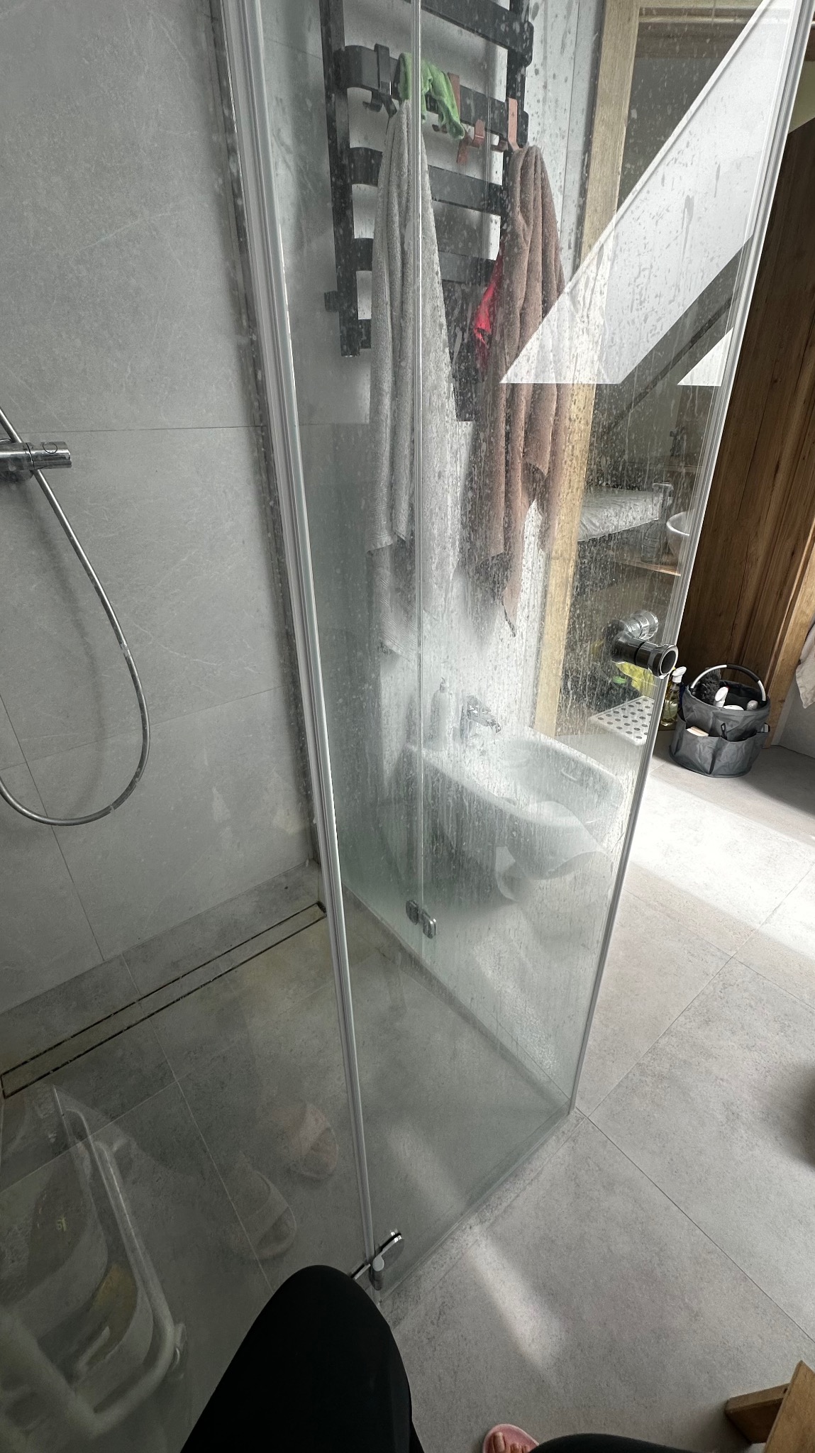 Brudna kabina prysznicowa z widocznymi zaciekami i osadami, z boku widoczny grzejnik łazienkowy z ręcznikami. Na podłodze widoczne klapki.
