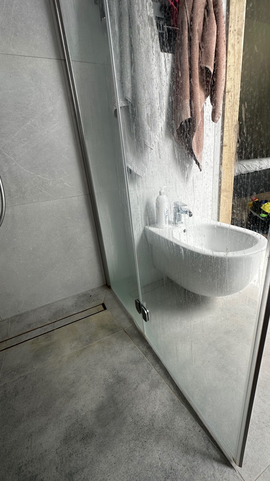 Zabrudzona kabina prysznicowa z bidetem i ręcznikiem w łazience z szarymi płytkami. Widoczne ślady po mydle i zaciekach. Potrzeba sprzątania po remoncie.