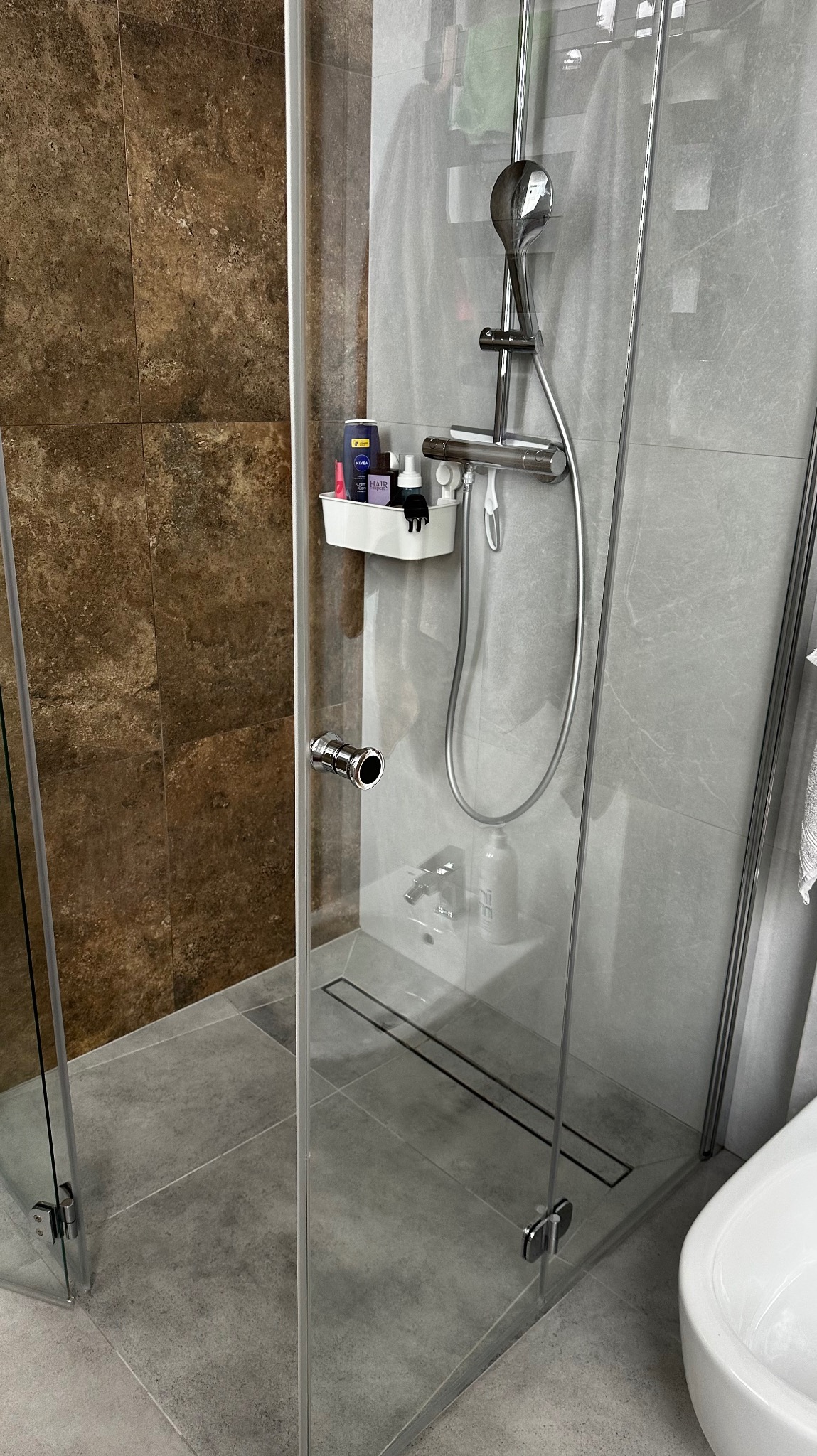 Nowoczesna kabina prysznicowa z odpływem liniowym, szklanymi drzwiami i półką na kosmetyki. Widoczne brązowe i szare płytki oraz chromowana armatura.