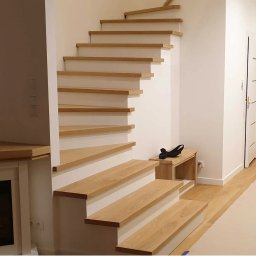 STOLAR Damian Krzysztof - Białe schody ze stopniami z jasnego drewna, bez poręczy, z małą drewnianą ławeczką pod spodem, w nowoczesnym jasnym wnętrzu z kominkiem i białymi drzwiami.