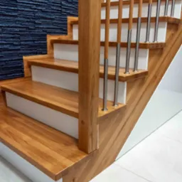 Drewniane schody z białymi podstopnicami i metalowymi balustradami na tle ściany z dekoracyjnej, ciemnej cegły. Widok z dołu, oświetlenie naturalne.