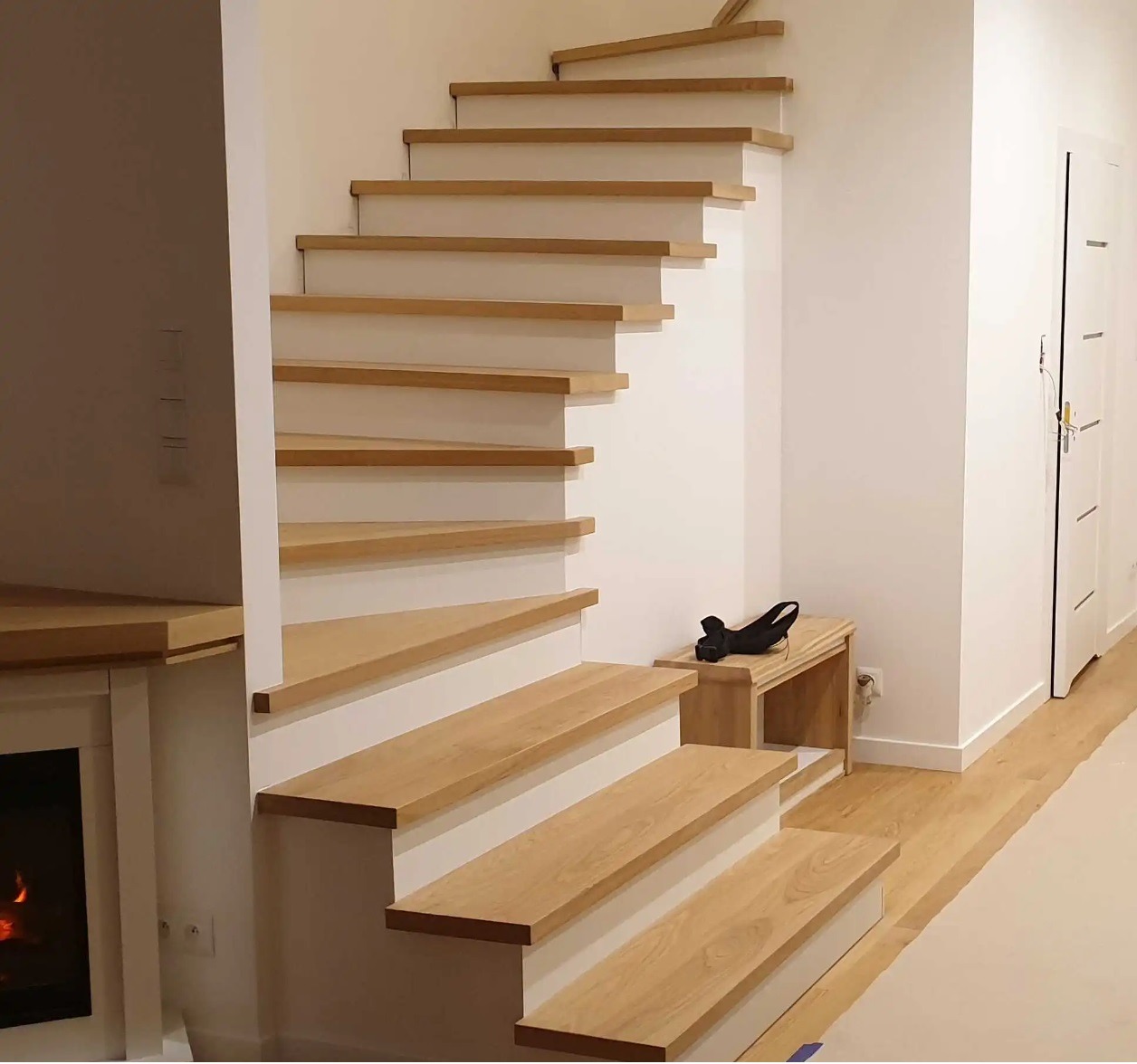 Białe schody ze stopniami z jasnego drewna, bez poręczy, z małą drewnianą ławeczką pod spodem, w nowoczesnym jasnym wnętrzu z kominkiem i białymi drzwiami.