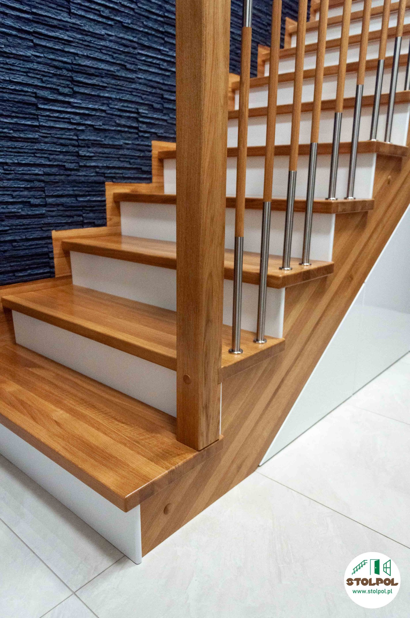 Drewniane schody z białymi podstopnicami i metalowymi balustradami na tle ściany z dekoracyjnej, ciemnej cegły. Widok z dołu, oświetlenie naturalne.