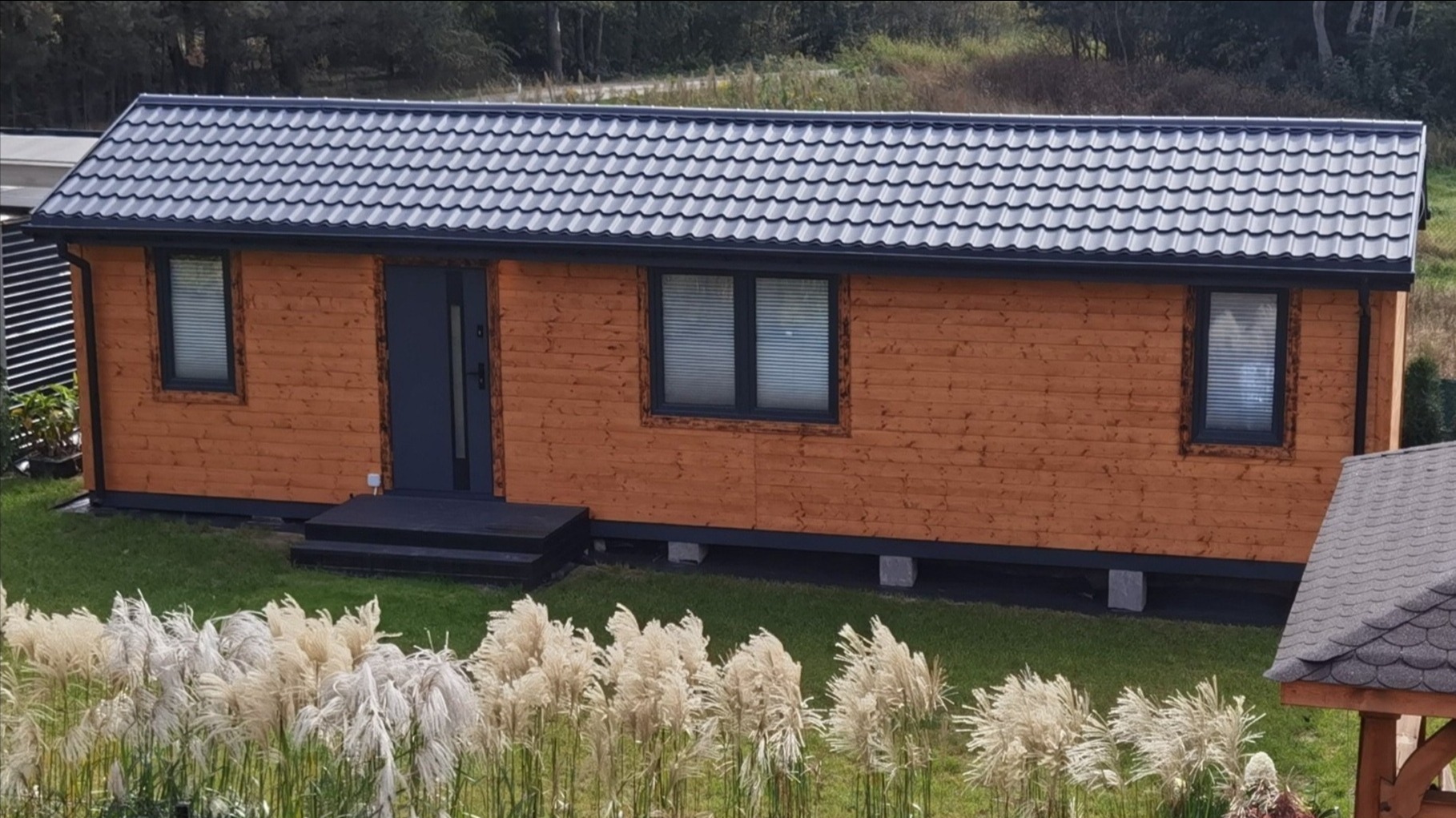 Gotowy, parterowy dom modułowy z drewnianą elewacją, szarą stolarką i dachem z blachodachówki, otoczony zielenią i ozdobnymi trawami.