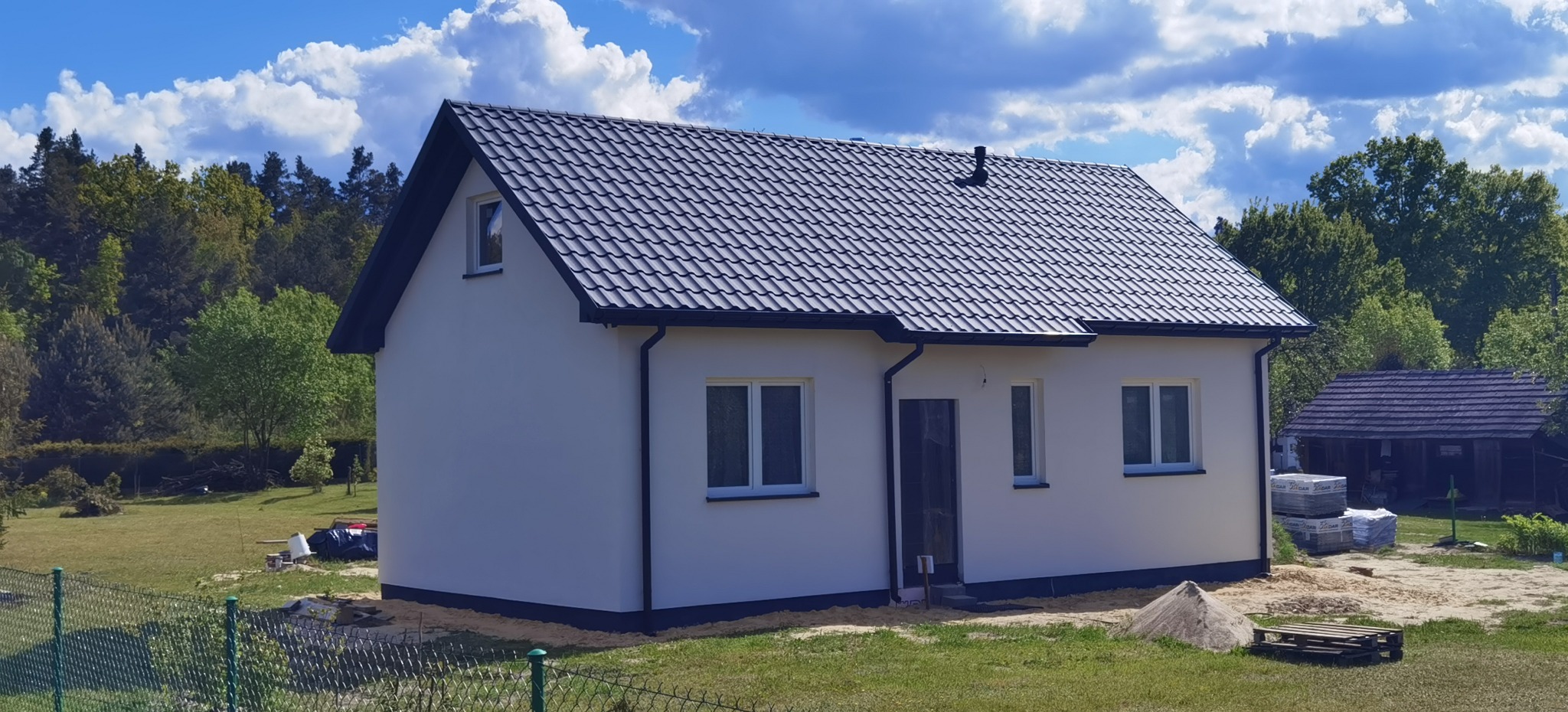 Biały, parterowy dom z ciemną dachówką na tle zieleni i błękitnego nieba z chmurami. Widoczne okna, drzwi wejściowe i elementy wykończeniowe.