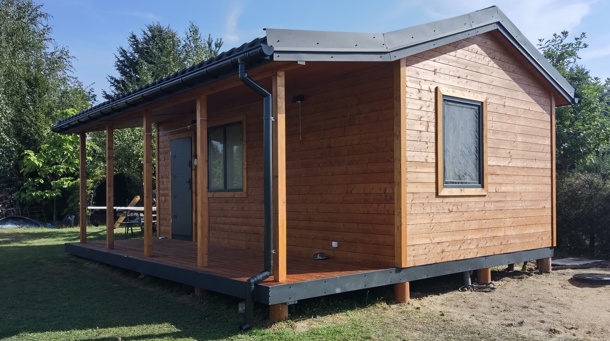 Drewniany domek modułowy z zadaszonym tarasem, podniesiony na betonowych słupach. Widoczne okno, drzwi i system rynnowy. Otoczenie: trawa i drzewa.