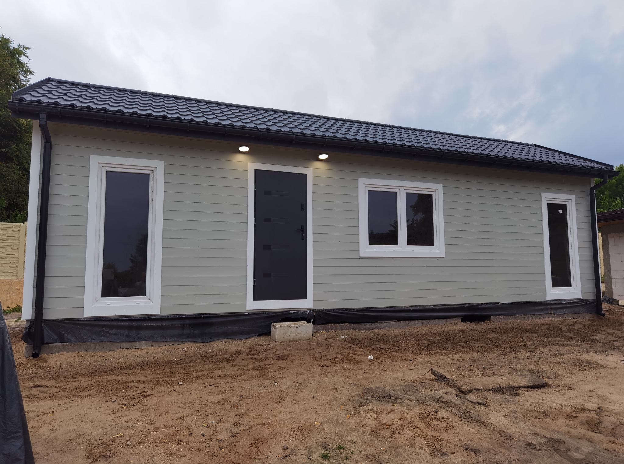 Gotowy, parterowy dom modułowy z szarą elewacją, czarnym dachem i stolarką okienną w kolorze białym. Dom stoi na działce z ziemią. Widoczne oświetlenie zewnętrzne.