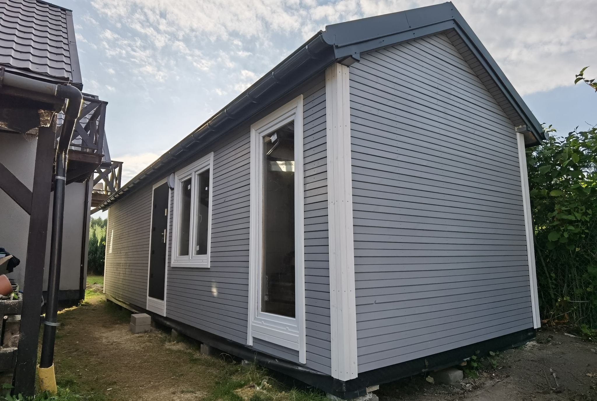 Gotowy, szary dom modułowy z białymi oknami w Lublinie. Widoczne poziome deski elewacyjne i fragment sąsiedniego budynku. Słoneczny dzień, błękitne niebo.