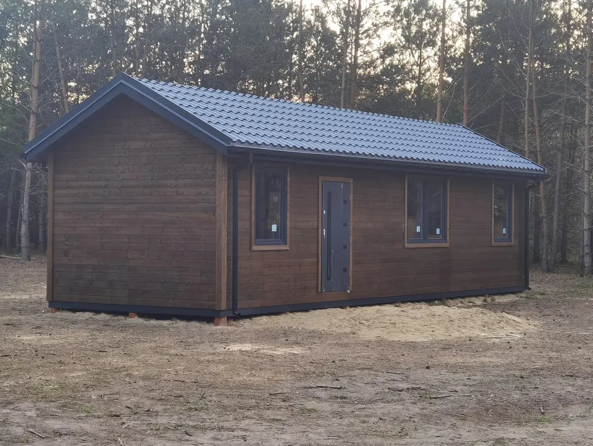 Gotowy, brązowy dom modułowy z ciemnoszarą stolarką i dachem z blachy na tle lasu iglastego. Budynek parterowy, z widocznymi oknami i drzwiami wejściowymi.