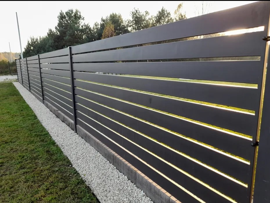 Nowoczesne, poziome ogrodzenie panelowe w kolorze antracytowym, z minimalistyczną podsypką z białego kamienia. Widok na tle zielonego trawnika i drzew.