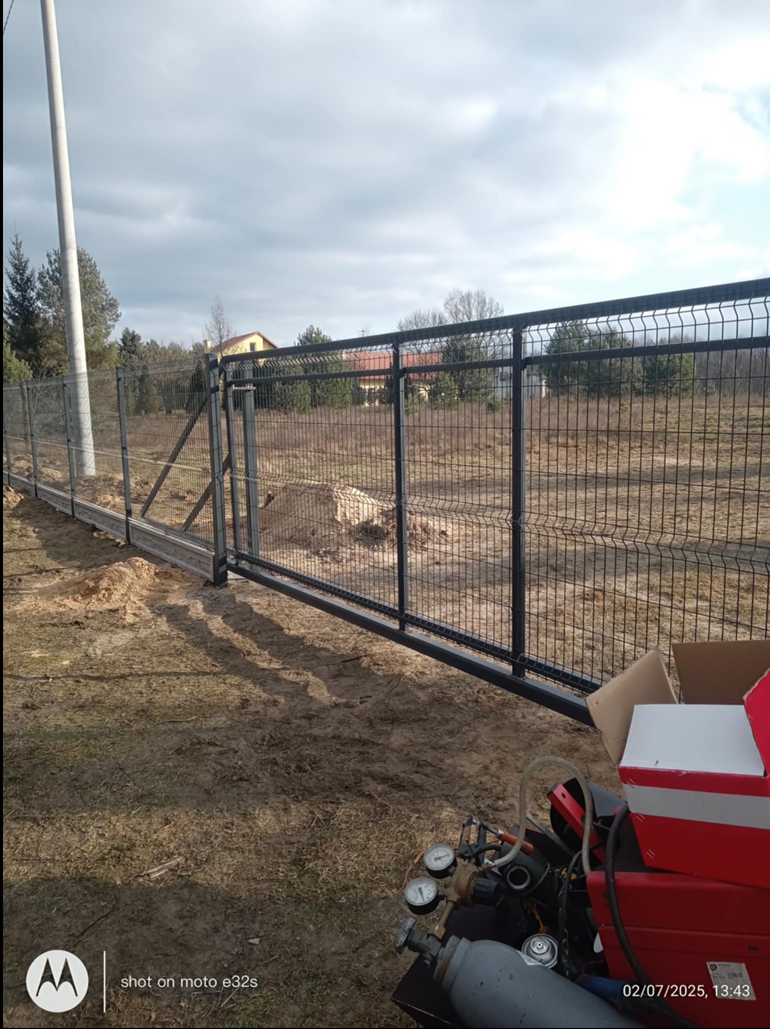 Nowoczesne ogrodzenie panelowe z bramą przesuwną, widoczne na tle działki z domem. W kadrze fragment sprzętu spawalniczego, sugerujący etap montażu.