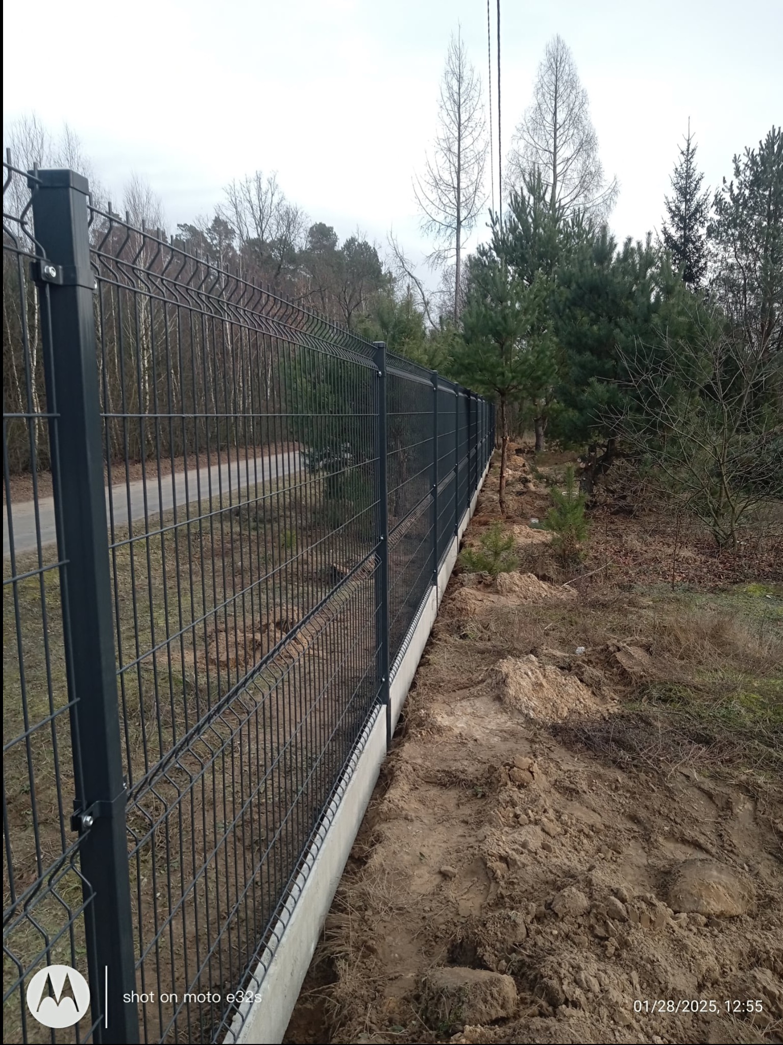 Długi, grafitowy panelowy płot na betonowej podmurówce, wzdłuż polnej drogi, otoczony drzewami. Widoczne ślady prac ziemnych przy płocie.